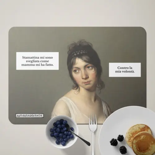 Tovaglietta americana Antipatiagratuita apparecchiata per la colazione, con una ciotola di mirtilli, una forchetta, un piatto di pancake con more e un tovagliolo bianco. Sopra, il ritratto di una giovane donna d&rsquo;epoca dall&rsquo;aria scocciata con i fumetti &ldquo;Stamattina mi sono svegliata come mamma mi ha fatto.&rdquo; e &ldquo;Contro la mia volont&agrave;.&rdquo;