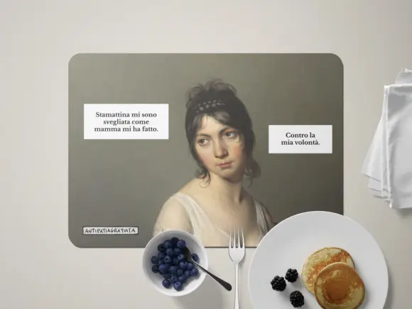 Tovaglietta americana Antipatiagratuita apparecchiata per la colazione, con una ciotola di mirtilli, una forchetta, un piatto di pancake con more e un tovagliolo bianco. Sopra, il ritratto di una giovane donna d&rsquo;epoca dall&rsquo;aria scocciata con i fumetti &ldquo;Stamattina mi sono svegliata come mamma mi ha fatto.&rdquo; e &ldquo;Contro la mia volont&agrave;.&rdquo;