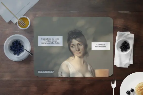 Tovaglietta americana Antipatiagratuita su un tavolo di legno scuro, circondata da miele, mirtilli, more, una forchetta e pancake. Sopra, il ritratto di una giovane donna d&rsquo;epoca dall&rsquo;aria scocciata con i fumetti &ldquo;Stamattina mi sono svegliata come mamma mi ha fatto.&rdquo; e &ldquo;Contro la mia volont&agrave;.&rdquo;