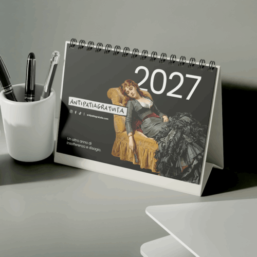 Calendario da tavolo 2027 Antipatiagratuita su una scrivania grigia, accanto a un portapenne bianco con tre penne e a una piccola pianta grassa. In copertina il dipinto di una donna annoiata su una poltrona dorata e la frase &ldquo;Un altro anno di insofferenza e disagio.&rdquo;