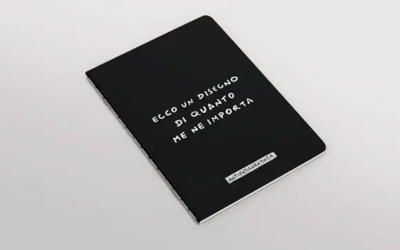 Quaderno nero con angoli arrotondati e cucitura a vista, inclinato in prospettiva su sfondo grigio chiaro. In copertina la frase scritta a mano in bianco &ldquo;Ecco un disegno di quanto me ne importa&rdquo; e, in basso, l&rsquo;etichetta &ldquo;Antipatiagratuita&rdquo;.