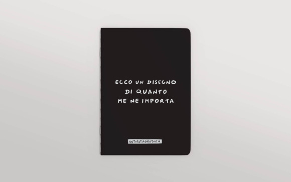 Quaderno nero con angoli arrotondati visto frontalmente su sfondo grigio chiaro. Sulla copertina, in bianco e in stile scritto a mano, la frase “Ecco un disegno di quanto me ne importa” e in basso l’etichetta “Antipatiagratuita”.