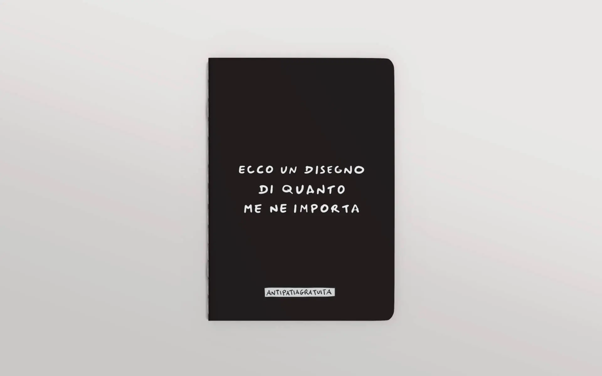 Quaderno nero con angoli arrotondati visto frontalmente su sfondo grigio chiaro. Sulla copertina, in bianco e in stile scritto a mano, la frase &ldquo;Ecco un disegno di quanto me ne importa&rdquo; e in basso l&rsquo;etichetta &ldquo;Antipatiagratuita&rdquo;.